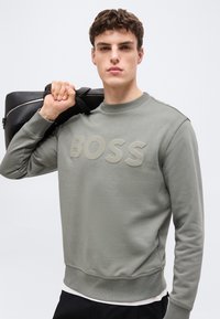 Νέος άνδρας που φοράει γκρι φούτερ με την επιγραφή «BOSS», κρατώντας μαύρη δερμάτινη τσάντα στον ώμο, κοιτάζει μπροστά με ουδέτερη έκφραση.