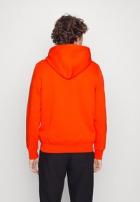 Oranje capuchontrui met een gestructureerd oppervlak, voorzien van een kangoeroezak, geribbelde manchetten en zoom. Achteraanzicht.