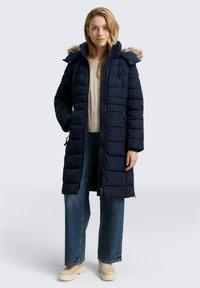 Eine Frau trägt einen langen marineblauen steppenden Mantel mit einer mit Pelz besetzten Kapuze, einen beigen Pullover, weite Jeans und beige Stiefel und steht mit einer Hand in der Tasche.