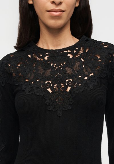Γυναίκα που φοράει μαύρο ριμπ τοπ με περίτεχνη δαντέλα με λουλουδένια μοτίβα γύρω από το λαιμό και το πάνω μέρος του στήθους, με απλό φόντο.
