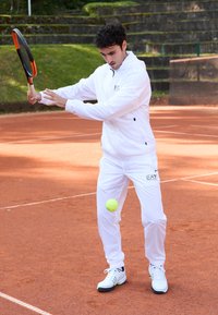 Valge spordirõivastus koos logoga, hoides tennisereketit, valmis lööma kollast tennisepalli punasel savikohtumisel. Roheline taust.