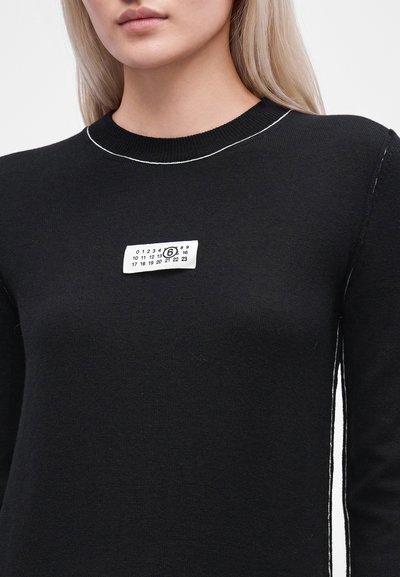 Personne blonde portant un pull noir avec des coutures blanches et un petit patch rectangulaire blanc montrant des chiffres, avec le chiffre six entouré sur la poitrine.