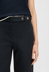 Schwarze hoch taillierte Hose mit glatter Textur und kontrastierendem weißen Besatz am Bund, der mit metallischen Druckknöpfen gesichert ist.
