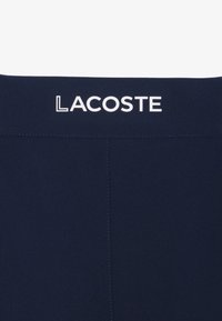 Tela azul oscuro con costura cosida y logo de texto blanco "LACOSTE" centrado cerca del borde superior.