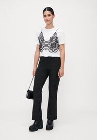 Jeune femme se tient debout, portant un haut blanc à manches courtes avec un superposition de dentelle noire, un pantalon à rayures noires scintillantes, des bottes à plateforme, tenant un petit sac à main noir.