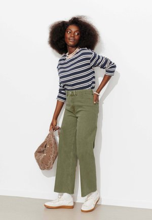 Femme aux cheveux bouclés et volumineux portant un shirt à manches longues rayé, un pantalon taille haute couleur olive, des baskets blanches, et tenant un sac marron.