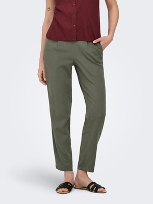 Vrouw die een olijfgroene broek met rechte pijpen draagt, een bordeauxrode mouwloze top met knopen en zwarte sandalen met open teen, staand met één hand in haar zak.