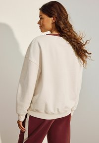 Sudadera beige clara de corte holgado, con hombros caídos y puños de canalé. Combinada con pantalones burdeos que presentan franjas laterales blancas.