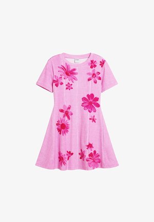 Ružové šaty s kvetinovým vzorom, krátkymi rukávmi a voľným strihom. Na ramene je prehozená ružová rifľová bunda v rovnakom odtieni. Strieborné sandále.