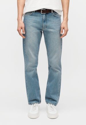 Férfi világoskék egyenes szárú farmernadrágban, fehér tornacipőben, fehér ingben és barna fonott övvel, egyszínű háttér előtt.