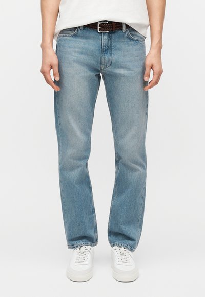 Homme portant un jean droit bleu clair, des baskets blanches, une chemise blanche et une ceinture tressée marron sur un fond uni.