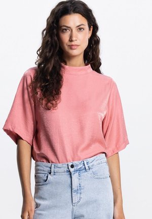 Vrouw met lang, krullend donker haar die een loszittend roze kort mouwtopje draagt, gestopt in een hoog getailleerde lichtblauwe spijkerbroek, naar voren kijkend.