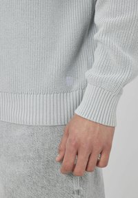 Un pull en tricot gris clair avec des poignets et un ourlet côtelés, orné d'une discrète étiquette logo sur le côté, porté sur un pantalon gris texturé.