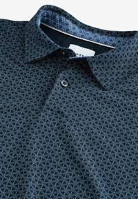 Námořnická modrá polo košile vyrobená z texturované látky má geometrický vzor ve světle modré, límec a zapínání na knoflíky.