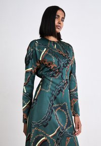 Langärmliges Kleid aus grüner Satin mit einem Kettenmuster in Braun, Schwarz und goldenen Akzenten. Verfügt über einen runden Ausschnitt und eine taillierte Passform.