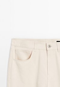 Pantalon beige taille haute avec poches latérales avant, passants pour ceinture et fermeture par un bouton argenté au niveau de la taille.