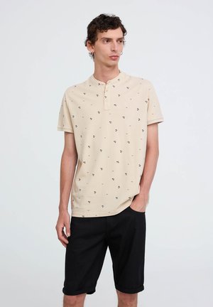 Polo beige a maniche corte con colletto bottonato e piccoli motivi floreali neri, abbinato a pantaloni corti neri arrotolati.