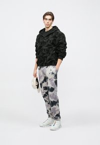 Melna rāvējslēdzēja kapuce ar teksturētu dizainu, kopā ar pelēkām kaklasaites krāsas biksēm un baltiem sporta apaviem. Modelis tur bēša cepuri.