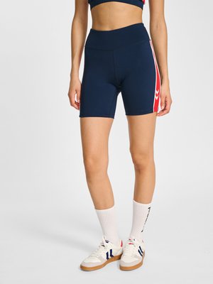 Kobieta nosząca granatowe sportowe szorty z wysokim stanem z czerwonymi i białymi bocznymi paskami, białe skarpetki oraz białe tenisówki z niebieskimi detalami.