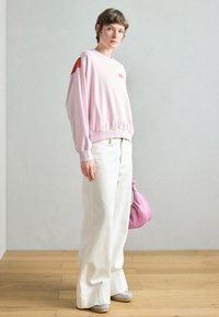 Svetloružová mikina so červenými akcentmi na ramenách, biele široké nohavice a sivé topánky. Modelka drží ružovú kabelku na neutrálnom pozadí.