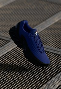 Blue athletic shoe with a textured upper, black sole, and distinctive air cushioning pods. Features rounded toe and lace-up design.