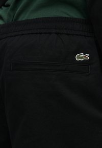 Πίσω τσέπη μαύρου παντελονιού με λαστιχένια μέση και πράσινο λογότυπο κροκοδείλου Lacoste στην πάνω δεξιά πλευρά.