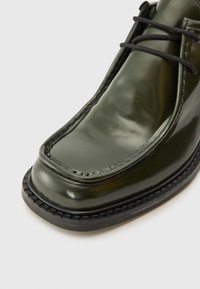 Green patent leather shoe with a rounded toe, black lace-up design, and textured black sole. Smooth finish with stitched detailing.