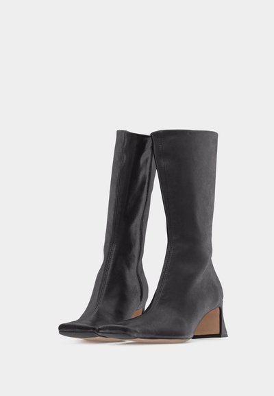 Ζευγάρι μαύρων δερμάτινων μπότες μέσης-γαστρίδας με τετράγωνα δάχτυλα και χαμηλά τακούνια μπλοκ, τοποθετημένες δίπλα-δίπλα σε λευκό φόντο.