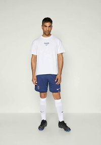 Balta Nike T-krekls ar uzrakstu "PARIS", zilas šortus ar logo, baltas līdz ceļiem zeķes un melnas futbola kurpes ar zilām akcentiem.
