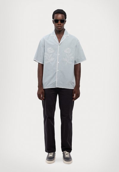 Šviesiai mėlyna trumpomis rankovėmis marškinėliai su balta gėlių siuvinėjimu, sagstomi priekyje, su kontrastinga siuvinėjimo spalva, derinami su tamsiomis kelnėmis ir pilkais sportiniais bateliais.