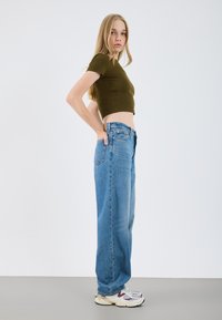 Vaaleanoliivinvihreä raidallinen crop-top yhdistettynä löysiin, korkeavyötäröisiin vaaleansinisiin farkkuihin. Mallilla on päällään valkoiset ja violetit lenkkarit. Yksinkertainen, minimalistinen tausta.