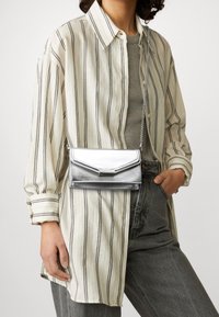 Srebrna metaliczna torebka na ramię o eleganckim designie, z łańcuszkowym paskiem i klapką typu fold-over, w zestawieniu z paskowaną koszulą na guziki i szarymi spodniami.