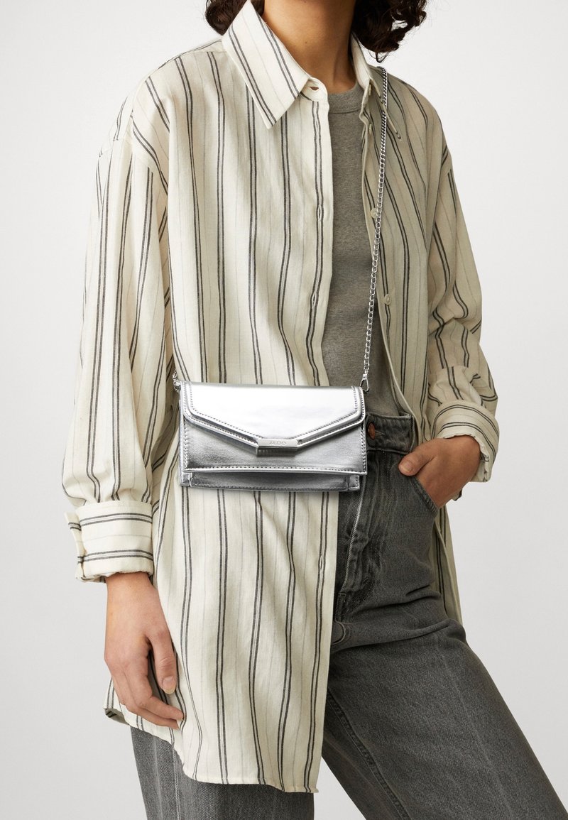 Srebrna metaliczna torebka na ramię o eleganckim designie, z łańcuszkowym paskiem i klapką typu fold-over, w zestawieniu z paskowaną koszulą na guziki i szarymi spodniami.