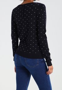 Granatowy sweter w białe kropki, z długimi rękawami i subtelnym guzikiem z tyłu. Tkanina wydaje się miękka i dopasowana. Noszony z niebieskimi dżinsami.