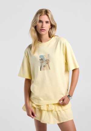 Jonge vrouw met blond haar, die een gele "Santa Monica" T-shirt en een bijpassende gele roezelrok draagt tegen een effen achtergrond.