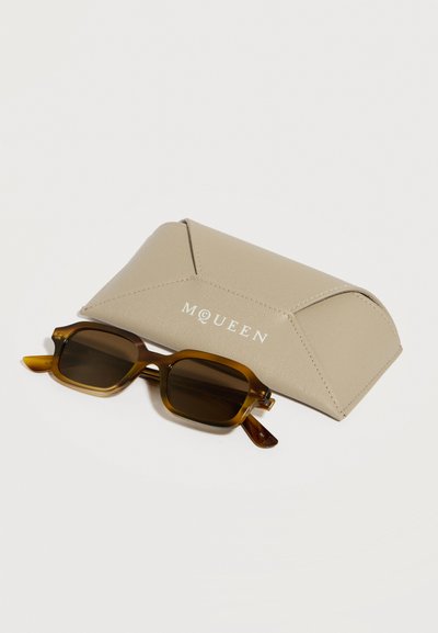 Ορθογώνια καφέ γυαλιά ηλίου με σκελετό από χελώνα, τοποθετημένα μπροστά από σκληρή θήκη McQueen σε μπεζ χρώμα, πάνω σε λευκό φόντο.