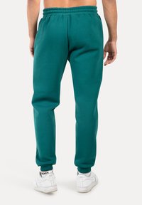 Tyrkysové tepláky s elastickým pasem a manžetami na kotnících, vyrobené z bavlněného materiálu, v kombinaci s bílými sportovními botami.