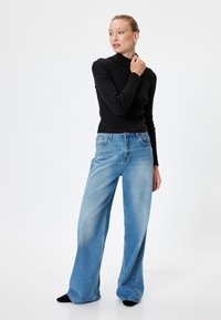Maglione nero a coste con collo alto abbinato a jeans a gamba larga di un blu chiaro, caratterizzati da una vestibilità morbida e un leggero effetto sbiadito, indossato con calzature nere.