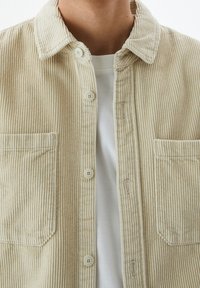 Chemise en velours côtelé beige avec col, fermeture à boutons et deux poches avant. Présente une texture côtelée verticale et des boutons crème.