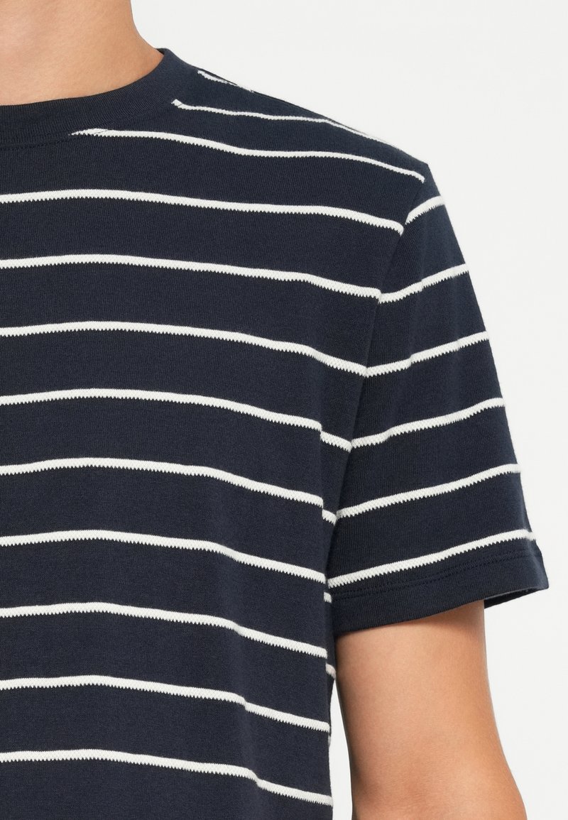 Granatowa koszulka z białymi poziomymi paskami, okrągłym dekoltem, krótkimi rękawami i miękką bawełnianą strukturą. Widoczne nitki przy dekolcie.