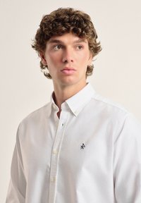 Camisa blanca de botones hecha de tejido texturizado, con un pequeño logo navy en el lado izquierdo. Cuello clásico y puños abotonados.