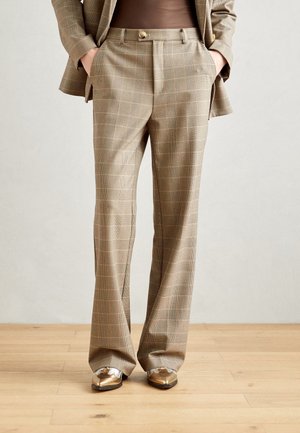 Persoon draagt beige geruite broek en bijpassend colbert met handen in de zakken, staand op een lichte houten vloer met metalen gouden schoenen.