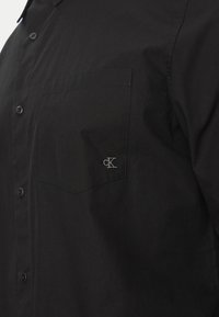 Camicia nera da uomo con bottoni, in tessuto di cotone, con una tasca sul petto e il logo "CK" ricamato in grigio sulla tasca. Texture liscia e design classico.