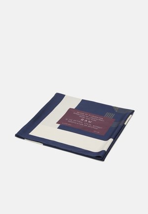 Foulard carré plié bleu marine et crème avec une étiquette bordeaux portant l'inscription « RAW » et un texte sur l'état naturel et la pureté.
