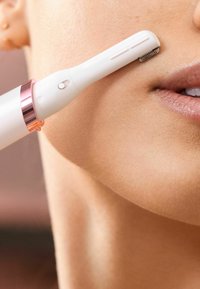Tondeuse faciale blanche avec un accent en or rose, au design mince et à la tête de coupe texturée, posée près de la peau lisse et des lèvres.
