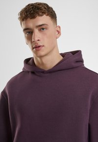 Lila Kapuzenpullover mit lässiger Passform, ausgestattet mit einer verstellbaren Kapuze und weichem Material. Das Design umfasst eine Kängurutasche und minimale Nähte.