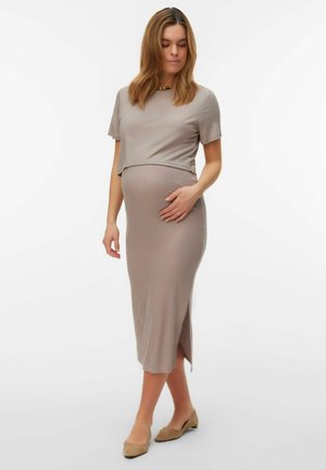 Nosečnica v bež dvodelni obleki z rebrastim vzorcem in kratkimi rokavi, ki drži trebuh, na nogah ima ravne bež čevlje, na belem ozadju.