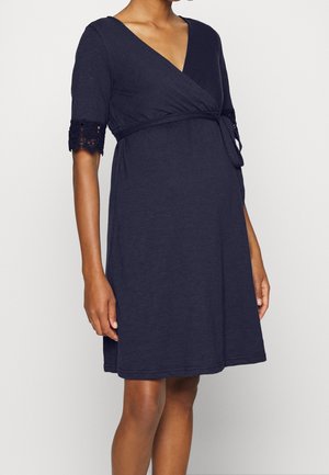 Těhotná osoba nosící tmavě modré zavinovací šaty po kolena s krajkovými detaily na rukávech délky ke loktům a zavázaným páskem v pase.