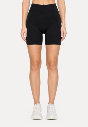 Mujer con shorts de ciclista negros de talle alto y zapatillas blancas, de pie frente a un fondo claro y liso.