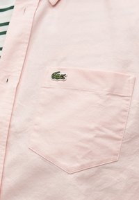 Poche sur une chemise rose clair à boutons, avec un petit logo de crocodile brodé en vert près du coin supérieur gauche.